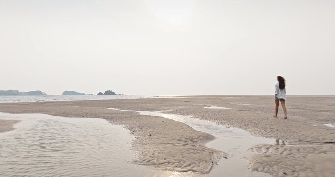 Woman walking barefoot across expansive tidal sandflats with shallow water pools reflecting the sky, revealing the natural beauty of the low tide landscape on a tropical beach in Koh Lanta. Back view