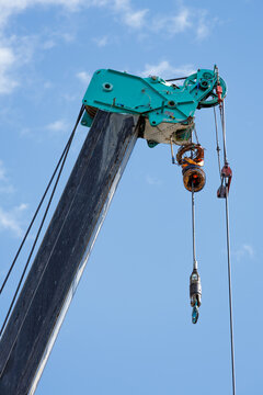 Extended heavy-duty crane boom with teal jib head showcases system of steel cables, pulleys, and suspended hoist hook ready for lifting. Set against clear blue sky backdrop.