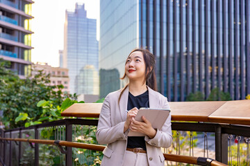 Luxury young Asian business woman entrepreneur using digital tablet while walking in the green building modern office downtown on background for communication and urban lifestyle