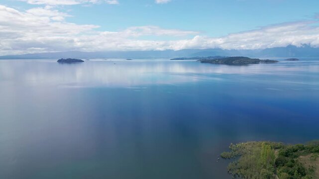 Forward flight over glassy lake surface with scattered islands and mountains. Lago Ranco, Chile