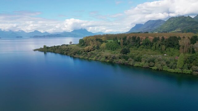 Orbit shot over forested peninsula with calm lake and distant Andean peaks. Lago Ranco, Chile