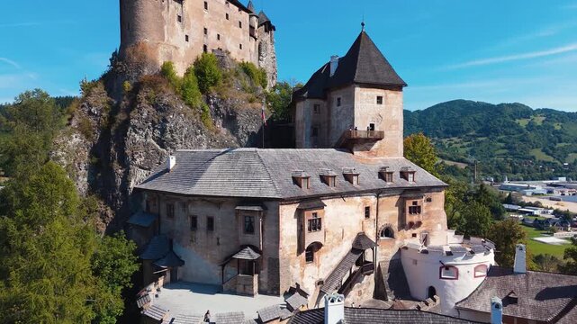 Drone pulling back from Orava Castle in Slovakia, revealing medieval stone buildings perched on dramatic rocky cliff above green valley.