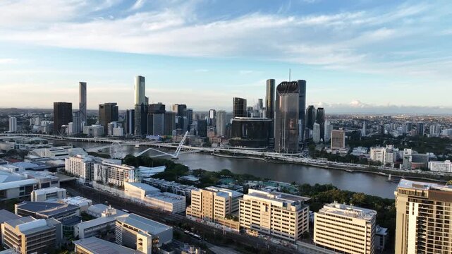 Slow drone side tracking at golden hour along the Brisbane River, holding Queens Wharf and The Star Brisbane towers with Neville Bonner Bridge and the CBD skyline, Brisbane, QLD, Australia