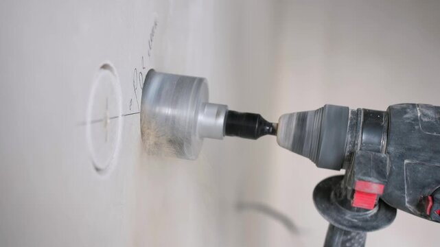 Worker drills circular openings in wall using hole saw for installing electrical socket boxes during renovation.