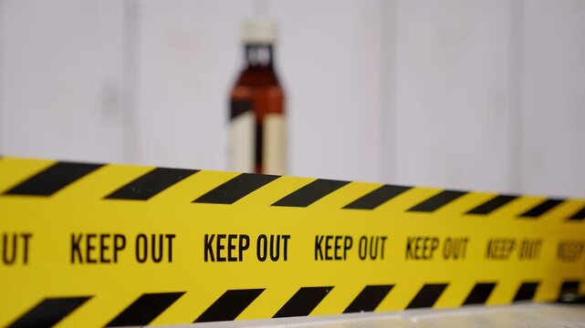Close-up of a yellow caution tape with the word KEEP OUT in the background, against the backdrop of a blurred bottle of alcohol. The issue of alcohol abuse.