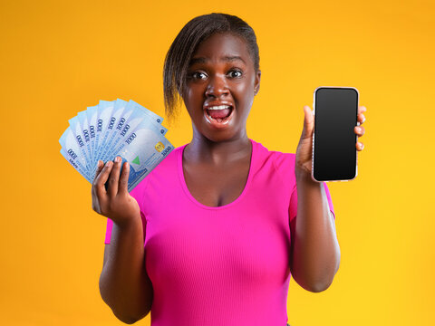 An excited young woman in a pink top poses against a yellow background. She joyfully displays a fan of new blue 1,000 Naira notes and holds a smartphone, symbolizing financial success or digital wealt