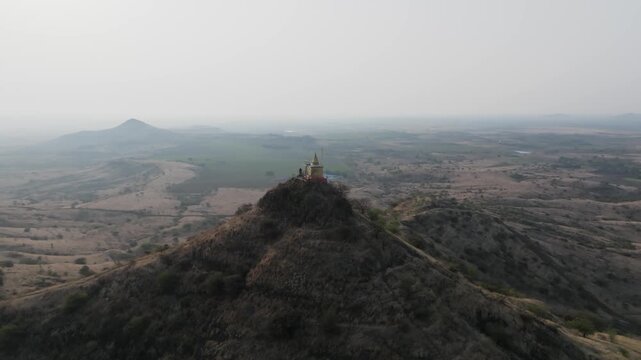 Cinematic drone orbits and pulls back from a Hindu temple perched on a dramatic rocky pinnacle revealing the vast hazy Deccan plateau near Garwad in Solapur Maharashtra