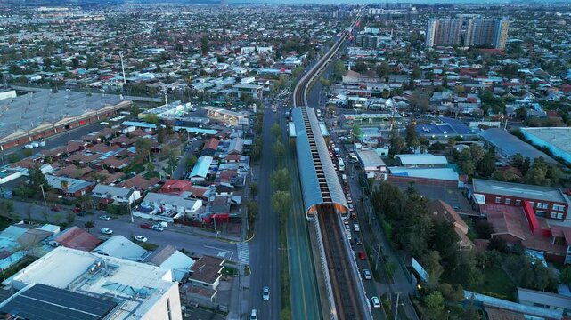 Aerial orbit shot around train with illuminated orange passenger cars on tracks cutting through urban residential area with heavy traffic on parallel highway at dusk. Santiago, Chile