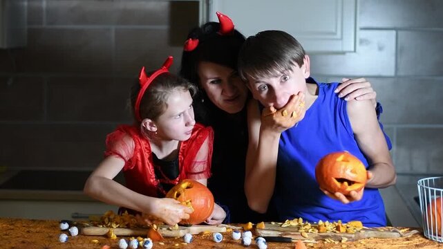 Happy family in devil costumes carving pumpkins together in the kitchen for Halloween party, with the son playfully eating pulp while preparing the spooky decorations at home