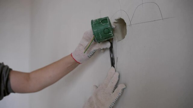 Electrician performs home renovation installing outlet box and laying wiring inside prepared wall groove. Apartment interior repair process.