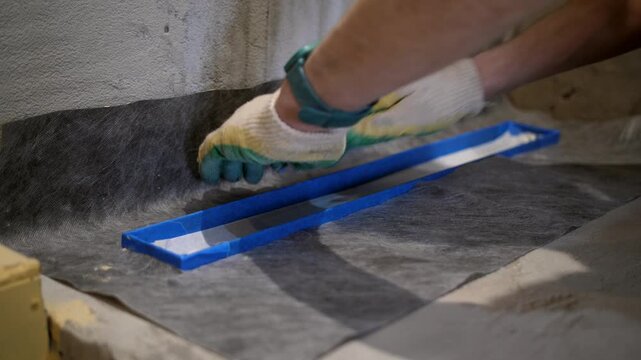 Close-up of shower drain covered with blue tape while worker installs waterproof underlay around the drain before applying liquid waterproofing. Bathroom renovation stage.