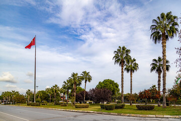 Obraz premium Turkish flag waving on a high flagpole along a city road with palm trees. Scenic urban landscape in Side, Antalya province, Turkey. Bright autumn day under a cloudy sky. Mediterranean.