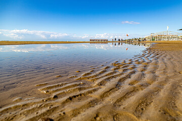 Low angle view of wavy sand structure and water puddle on a beach. Scenic coastal landscape with ripples, blue sky reflection, and a distant pier in Evrenseki, Side, Turkey. Autumn day.