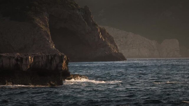 Golden hour light illuminating northern Ibiza cliffs as Mediterranean waves break against rocky outcrop creating calm yet dramatic coastal atmosphere.