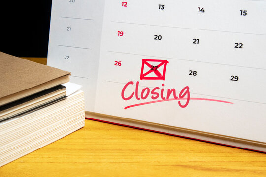 Close-up of a white desk calendar with the word Closing written in red ink and a specific date marked with a red X, placed on a wooden office desk next to a stack of notebooks.