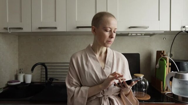 woman with short regrown hair after cancer treatment stands in a bright home kitchen wearing cozy loungewear and using a smartphone, relaxed female patient leaning on the countertop and texting online