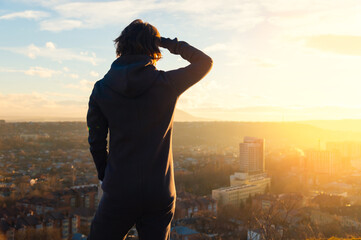 Female figure in a fleece jumpsuit stands on hilltop overlooking cityscape at sunset, with buildings and distant mountains visible under a colorful sky © yanik88