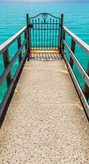 Fototapeta premium Perspective shot of concrete pier with wooden railing and metal gate overlooking turquoise sea, representing escape, travel and seaside destination
