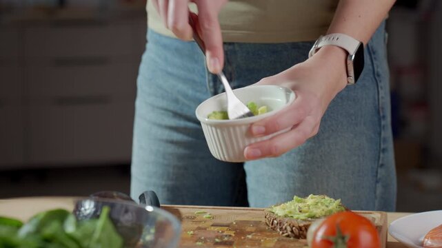 Woman mashing avocado with fork in white bowl close up. Concept of homemade guacamole preparation