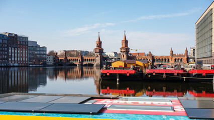 Red industrial river barges moored on the Spree River in the foreground contrast with a row of wooden market stalls and the iconic red-brick twin towers of the Oberbaumbrücke bridge