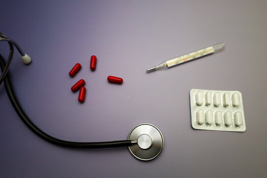 A professional medical still life featuring a black stethoscope, a glass thermometer, and various medications on a calm purple background. This composition is ideal for illustrating flu diagnostics, h