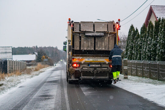Garbage truck collecting waste in winter snowfall, municipal worker riding on the back of a refuse vehicle, waste management during bad weather, snowy suburban road service.