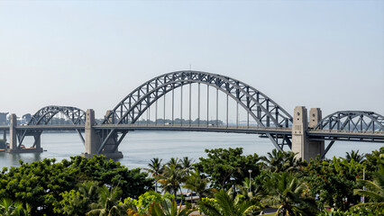 Fototapeta premium Large Steel Arch Bridge Spanning Wide River with Lush Green Palm Trees in Foreground under Clear Sky for Travel Architecture and Urban Development Concepts