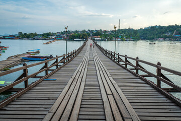 Obraz premium Scenic wooden Mon Bridge stretching across calm river toward local village at sunset in Sangkhlaburi, Kanchanaburi, Thailand