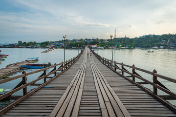 Fototapeta premium Scenic wooden Mon Bridge stretching across calm river toward local village at sunset in Sangkhlaburi, Kanchanaburi, Thailand