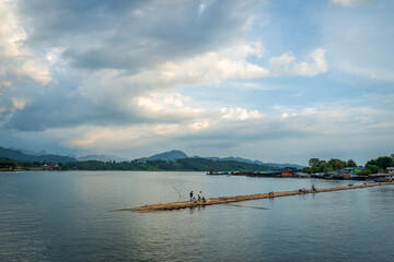 Fototapeta premium Scenic bamboo bridge crossing calm river with local village and mountain backdrop in Sangkhlaburi, Kanchanaburi, Thailand under dramatic cloudy sky