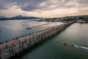 Naklejka premium Aerial sunset view of Mon Bridge over Songkalia River in Sangkhlaburi, Kanchanaburi, Thailand with longtail boat passing beneath the wooden bridge and dramatic cloud over tranquil water