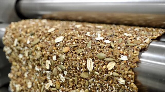 Closeup on multigrain dough layered with seeds and grains passing through laminating rollers to create textured nutritious cracker sheets on production line.