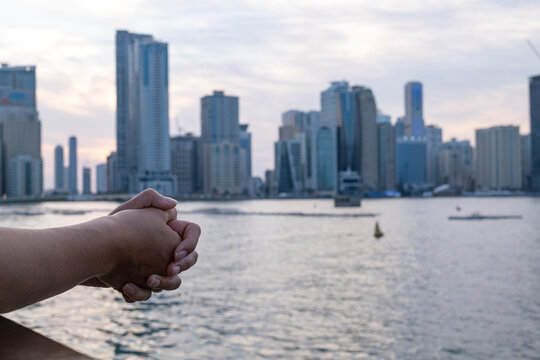 Close-up of hands leaning on waterfront railing with blurred Sharjah city skyline, skyscrapers and Khalid Lagoon in the background at sunset, UAE.