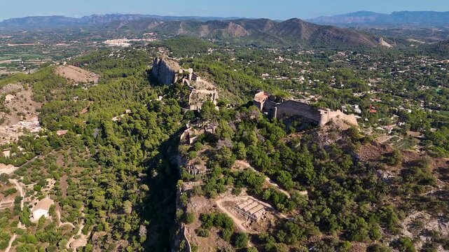 Aerial drone flying forward over the historic Xativa Castle on a mountain ridge in Valencia, Spain, featuring medieval walls and lush green surroundings.