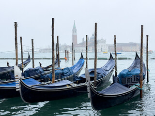 Gondolas moored in the water with wooden poles, foggy atmosphere, and historic buildings visible in the background in Venice, Italy
