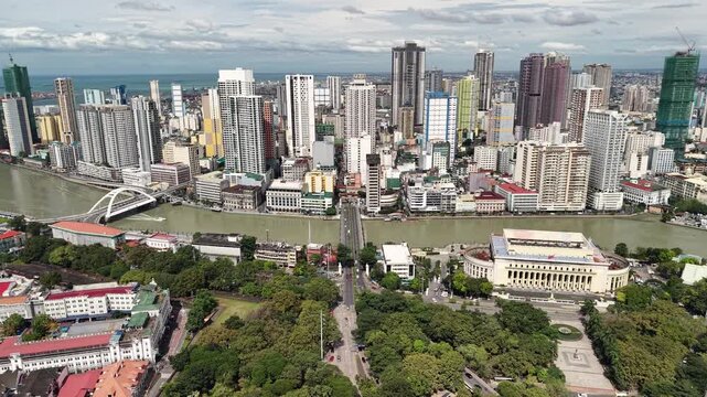 Wide elevated aerial captures Pasig River panorama from Intramuros park grounds showing white arch bridge and classical government building with towering modern Manila skyline fading into hazy horizon