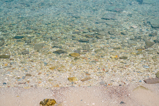 Clear turquoise ocean water gently flows over the pebbly bottom under bright sunlight, creating shimmering light patterns on the shallow shore, close-up