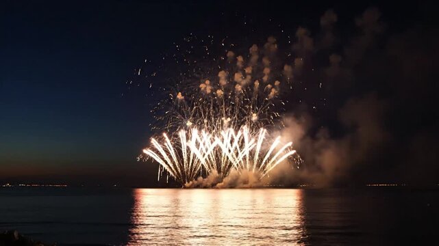 Spectacular golden and white fireworks explode in a dark night sky over a calm sea, reflecting their brilliant light on the water's surface.