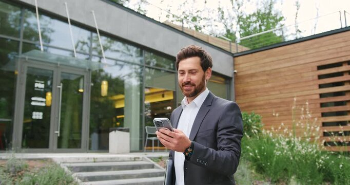 Confident businessman in a formal suit using smartphone for online communication while walking outdoors near modern office building
