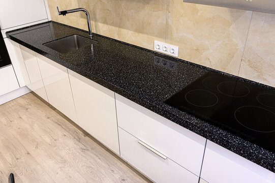A modern kitchen with a black speckled granite countertop, stainless steel sink, induction cooktop, white cabinetry, beige tiled backsplash, and light wood flooring