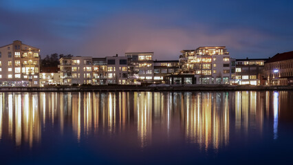 Obraz premium Authentic Scandinavian river culture illuminated residential waterfront in Porsgrunn Norway with glowing apartments mirrored in still river under northern evening sky highlighting local urban identity