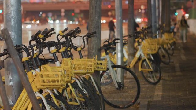 Night street lined with yellow bikes, wet pavement reflecting neon lights, rider locks rental bike near metal post while pedestrians pass and distant traffic hum creates gritty sharing economy mood