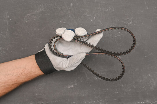 hand in a work protective glove of auto technician holding rubber belt in workshop. new timing belt, part of repair work and technical diagnostics. close-up on a grey concrete background
