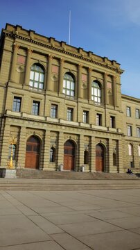 Vertical panning shot of the ETH Zurich main building in Switzerland, showcasing its grand 19th-century architecture and the spacious Polyterrasse plaza under a clear blue sky.