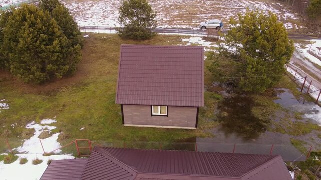 Aerial view of a Belarus homestead, brown roof cottage, greenhouse, fenced plots, muddy lane, striped fields of snow and growth, conifers, car and person, overcast light