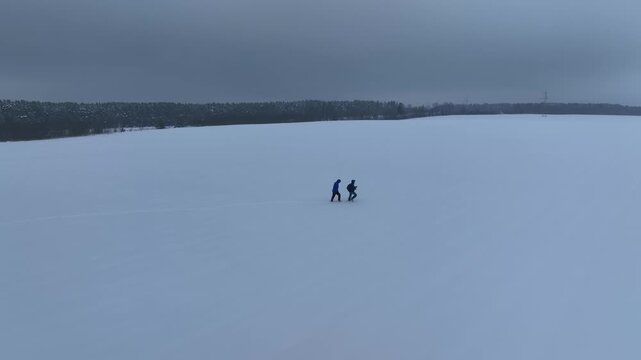 Aerial view shows two figures walking on a snowy field in rural Belarus, a dark coniferous tree line on the horizon, and a power pylon at right in muted light.