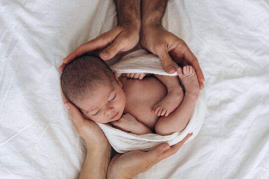 Newborn baby wrapped in soft cloth, gently cradled by two pairs of hands in peaceful sleep