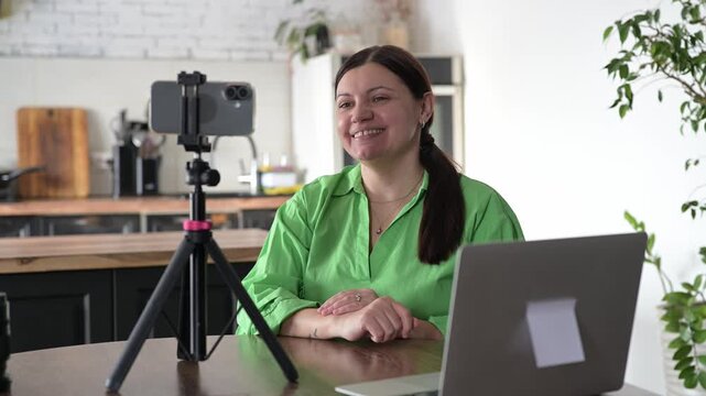 Female blogger aged 45 recording educational stream with smartphone on tripod in home interior. Content creation for online learning and knowledge sharing.