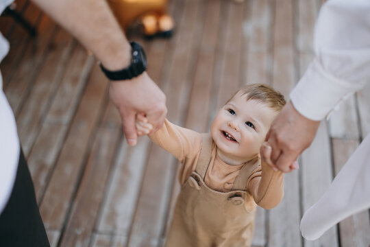Tiny child laughs while holding hands with two adults walking on wooden deck in warm sunny light