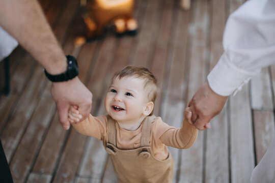 Tiny baby laughs with joy as two adults hold hands with her on wooden deck Tiny child laughs joyfully while holding hands with two adults on wooden deck in warm sunlight